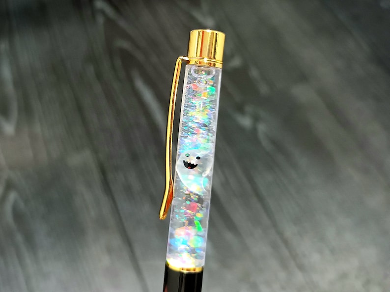 May include: A gold and clear pen with a smiling ghost design inside. The pen is filled with iridescent glitter.