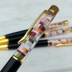 May include: A black and gold pen with a clear barrel filled with colorful confetti-like pieces. The pen has a gold clip and a gold cap.