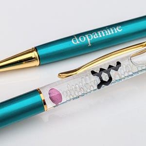 May include: Two pens with teal barrels and gold accents. The word "dopamine" is printed on the barrel of one pen. The other pen has a clear barrel with a black graphic and a pink shape inside. The pens are on a white surface.