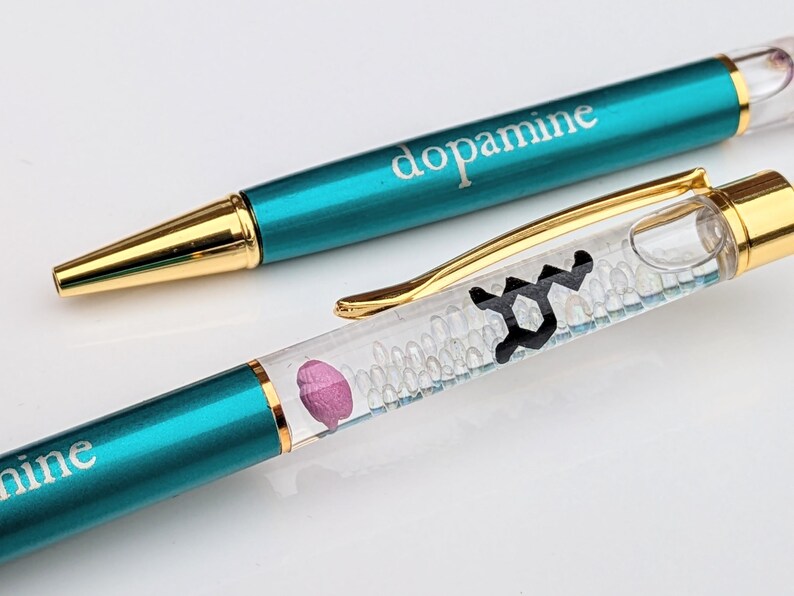 May include: Two pens with teal barrels and gold accents. The word "dopamine" is printed on the barrel of one pen. The other pen has a clear barrel with a black graphic and a pink shape inside. The pens are on a white surface.