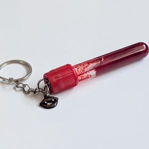 May include: A novelty keychain featuring a clear, cylindrical vial filled with a dark red liquid, resembling blood. The vial has a red cap and a small, dark charm shaped like vampire fangs. The keychain includes a silver keyring and chain.