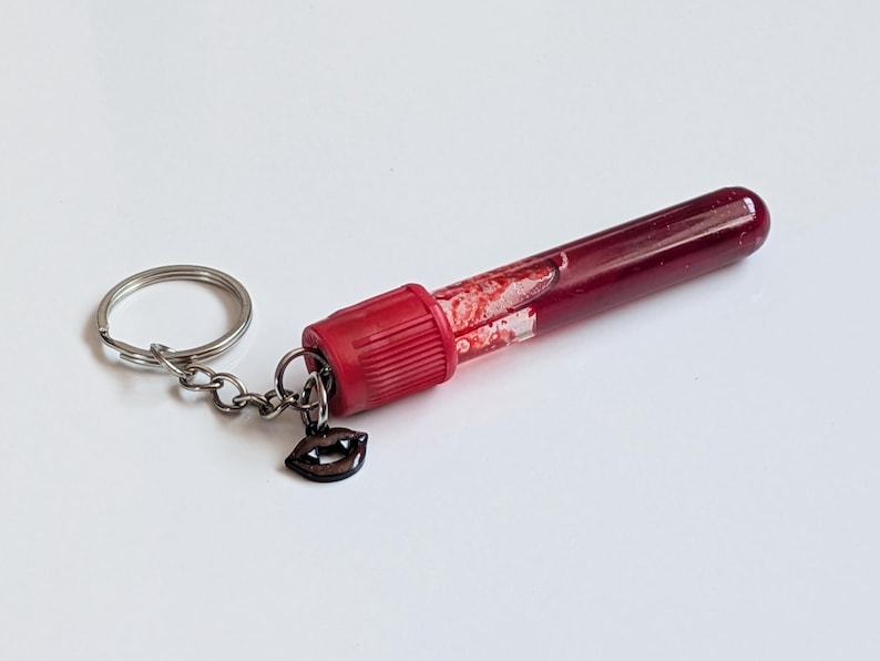 May include: A novelty keychain featuring a clear, cylindrical vial filled with a dark red liquid, resembling blood. The vial has a red cap and a small, dark charm shaped like vampire fangs. The keychain includes a silver keyring and chain.