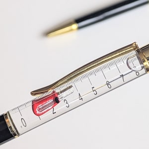 May include: Close-up of a pen with a clear barrel and a gold-colored clip and accents. The barrel has a numbered scale from 0 to 7. A second pen with a black barrel and gold accents is in the background.