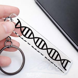 以下が含まれることがあります： 黒いDNA二重らせんのデザインが施された、透明な長方形のキーホルダー。キーホルダーは銀色のリングと小さな接続リングが付いています。背景には黒いノートがあります。