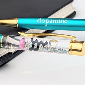 May include: Two pens with teal barrels and gold accents. One pen has the word "dopamine" printed on it. The other pen has a clear barrel with a pink brain, black molecule, and small iridescent beads inside.
