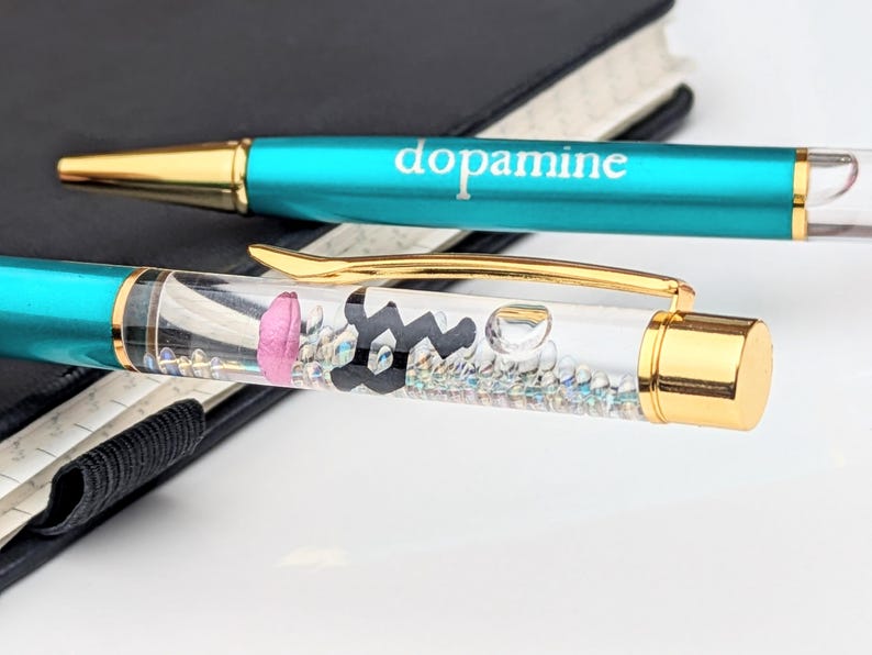 May include: Two pens with teal barrels and gold accents. One pen has the word "dopamine" printed on it. The other pen has a clear barrel with a pink brain, black molecule, and small iridescent beads inside.