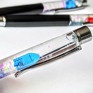 Könnte beinhalten: Ein transparenter Stift mit einer silbernen Kappe und einem blauen Bootsanhänger. Der Stift ist mit schimmerndem Glitzer und klarer Flüssigkeit gefüllt.