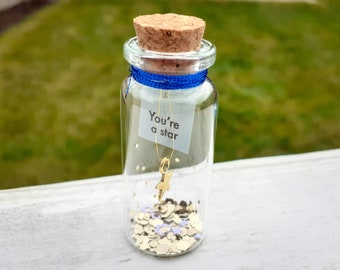 May include: A small glass jar with a cork lid, filled with gold and white glitter. A gold star charm hangs from a blue ribbon tied around the jar. A white paper tag with the text "You're a star" is attached to the ribbon.