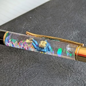 Puede incluir: Un bolígrafo negro y dorado con un cuerpo transparente lleno de purpurina de colores y una figura en miniatura de un buceador.