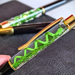 Op de afbeelding: Een transparante pen met een gouden dop en een groen slangontwerp erin. De pen ligt op een zwart oppervlak.