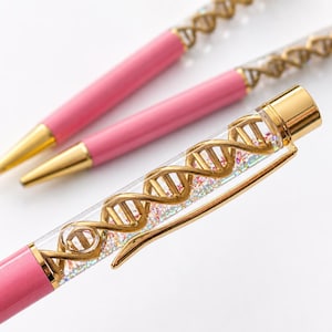 Puede incluir: Dos bolígrafos rosas con detalles en tono dorado. El cuerpo superior de cada bolígrafo es transparente y contiene una estructura de doble hélice de ADN de color dorado rodeada de pequeñas partículas de purpurina iridiscente y un clip dorado.