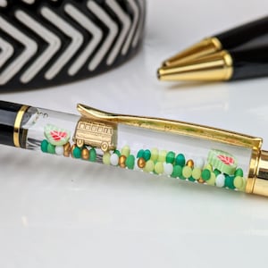 Könnte beinhalten: Dekorativer Stift mit einem transparenten Schaft, gefüllt mit grünen, weißen und gelben Perlen, einem goldfarbenen Bus-Anhänger und Wassermelonen-Scheiben-Anhängern. Der Stift hat einen schwarzen Körper und goldfarbene Akzente.