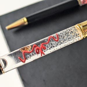 Op de afbeelding: Close-up van twee pennen. De primaire pen heeft een transparante behuizing gevuld met kleine belletjes en een rood, kronkelig ontwerp. De uiteinden van de pen zijn goudkleurig en de behuizing is zwart. De tweede pen is zwart met een goudkleurige punt.