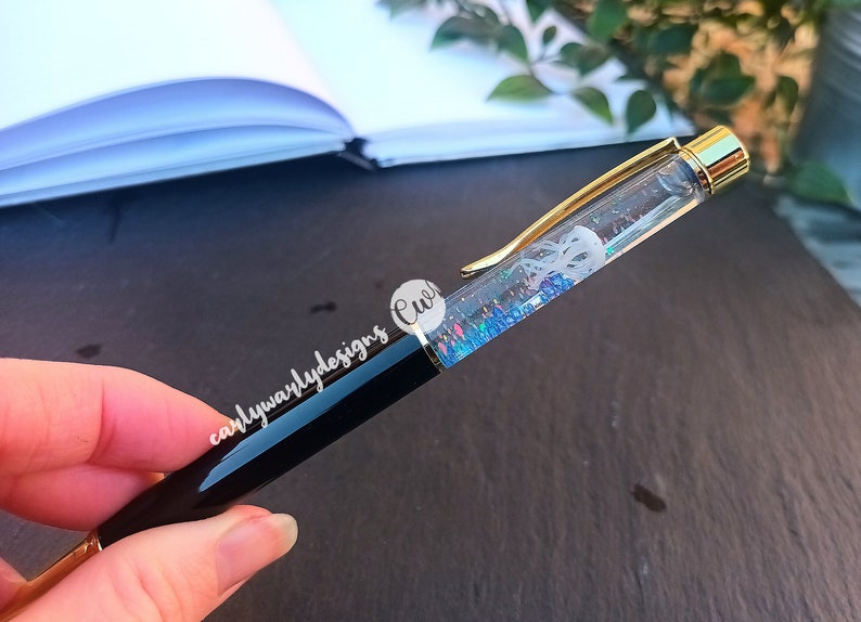 May include: A black and gold pen with a clear barrel filled with iridescent glitter and a white silhouette of a jellyfish. The pen is held in a hand.
