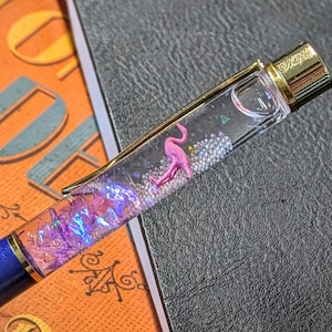 Może przedstawiać: Niebieski długopis z przezroczystym korpusem wypełnionym różowym flamingiem, srebrnymi koralikami i iryzującymi fioletowymi płatkami. Długopis ma złoty klips i końcówkę, leżący na teksturowanej powierzchni.