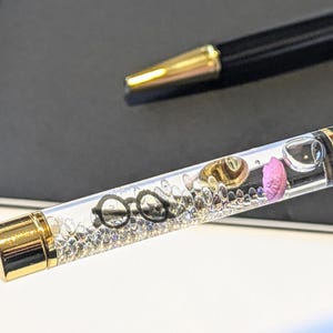 May include: Black and gold pen with a clear barrel filled with decorative elements, including eyeglasses, a gold-colored hat, and a pink object. The pen is set against a dark background, with another pen visible in the background.