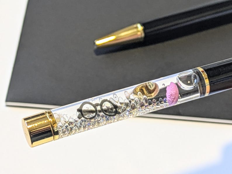 May include: Black and gold pen with a clear barrel filled with decorative elements, including eyeglasses, a gold-colored hat, and a pink object. The pen is set against a dark background, with another pen visible in the background.