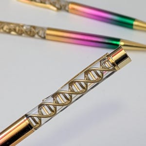 Könnte beinhalten: Ein goldener Stift mit einem durchsichtigen Schaft, der ein goldenes DNA-Helix-Design aufweist. Der Stift hat eine goldene Kappe und ein Farbverlaufsfinish, das von Pink zu Grün übergeht.