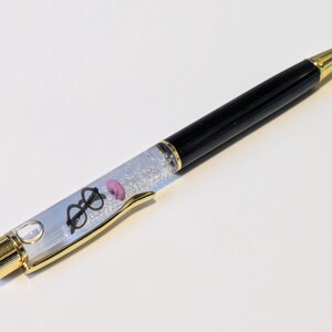 May include: A black and gold ballpoint pen with a clear barrel filled with glitter and small charms. The charms include a pair of glasses and a pink donut. The pen has a gold tip and accents.