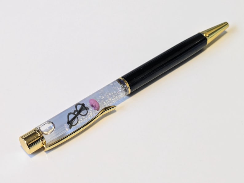 May include: A black and gold ballpoint pen with a clear barrel filled with glitter and small charms. The charms include a pair of glasses and a pink donut. The pen has a gold tip and accents.