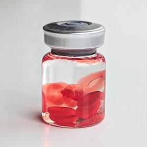 May include: A small, clear glass vial with a silver cap, filled with a red liquid and several red, oval-shaped objects. The vial is on a white surface.