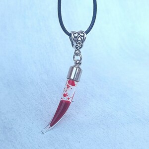 May include: A clear glass pendant necklace with a curved, horn-like shape filled with red liquid and red speckles. The pendant is suspended from a black cord and has a silver-colored metal cap and decorative bail.