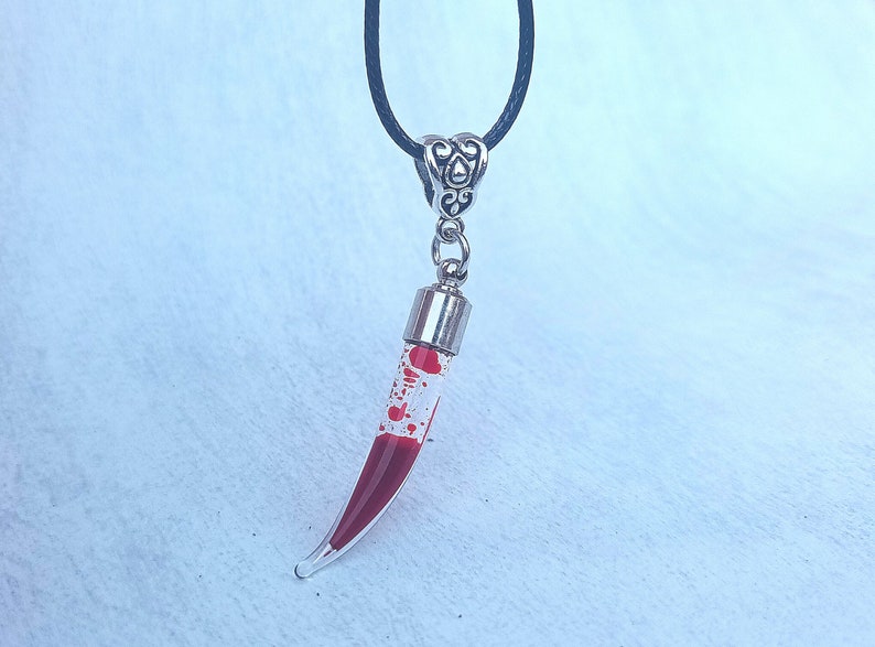 May include: A clear glass pendant necklace with a curved, horn-like shape filled with red liquid and red speckles. The pendant is suspended from a black cord and has a silver-colored metal cap and decorative bail.