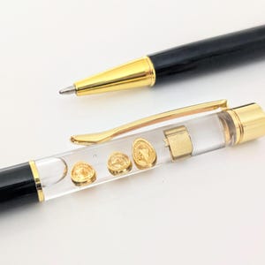 May include: A black ballpoint pen with a gold-toned tip and a clear acrylic barrel section containing three small gold-colored circular tokens and a rectangular gold-toned piece. The pen features a gold-toned pocket clip and matching end cap.
