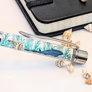 Peut inclure: Un stylo bleu sarcelle et argenté avec un corps transparent orné d'un dauphin bleu et de plantes aquatiques. Le stylo est entouré de coquillages et se trouve près d'un carnet noir.