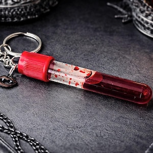 May include: A novelty keychain featuring a clear vial filled with red liquid, resembling blood, with red accents. The keychain includes a small charm of vampire fangs. The vial is attached to a silver keyring.