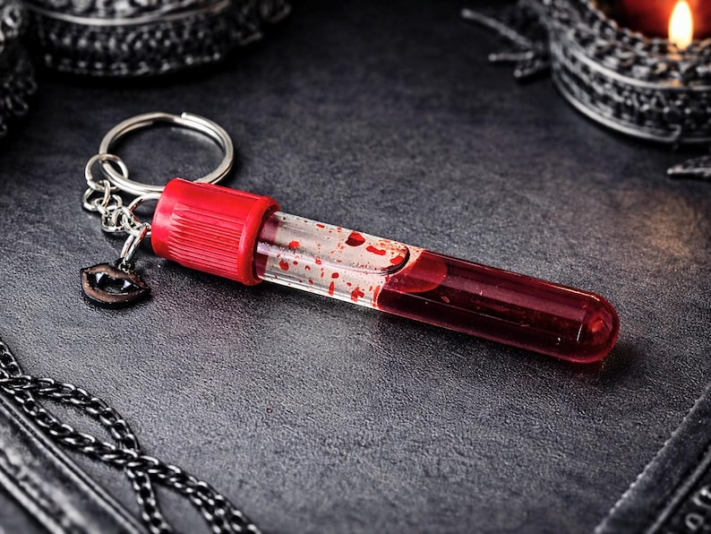 May include: A novelty keychain featuring a clear vial filled with red liquid, resembling blood, with red accents. The keychain includes a small charm of vampire fangs. The vial is attached to a silver keyring.