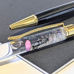 May include: A black and gold ballpoint pen with a clear barrel filled with decorative elements. The pen has a gold clip and tip, and a pink, flower-like object is visible inside the barrel. The pen is lying on a dark surface.