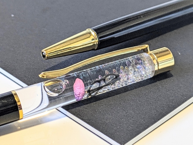 May include: A black and gold ballpoint pen with a clear barrel filled with decorative elements. The pen has a gold clip and tip, and a pink, flower-like object is visible inside the barrel. The pen is lying on a dark surface.
