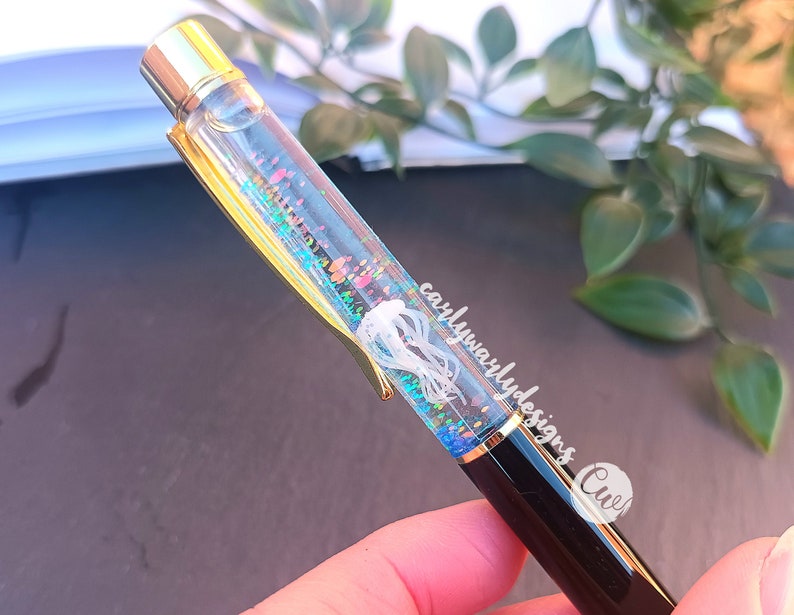 May include: A black and gold pen with a clear barrel filled with iridescent glitter and a white jellyfish. The pen has the text "carlywarlydesigns" and "Cute" written on it.