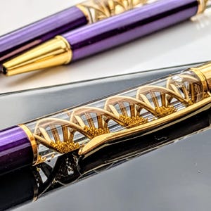 May include: A close-up shot of a purple and gold pen featuring a DNA helix design within a clear barrel. The pen has gold detailing and a gold nib. The background is a blurred black and white surface.
