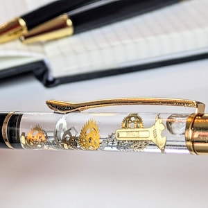 Könnte beinhalten: Nahaufnahme eines schwarz-goldenen Stifts mit einem transparenten Schaft, der mit Miniatur-Zahnrädern aus Gold und Silber und einem Schraubenschlüssel gefüllt ist. Der Stift hat einen goldenen Clip und eine goldene Spitze. Ein zweiter Stift ist im Hintergrund zu sehen.