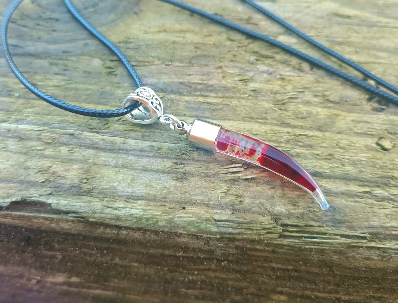 May include: A pendant necklace featuring a clear, curved horn-shaped charm filled with red and white inclusions. The charm is attached to a silver-colored metal cap and hangs from a black cord necklace. The background is a weathered wooden surface.