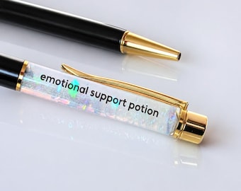 May include: Two pens on a white surface. One pen is black with gold accents. The other pen has a clear barrel filled with iridescent glitter and the words "emotional support potion" in black.