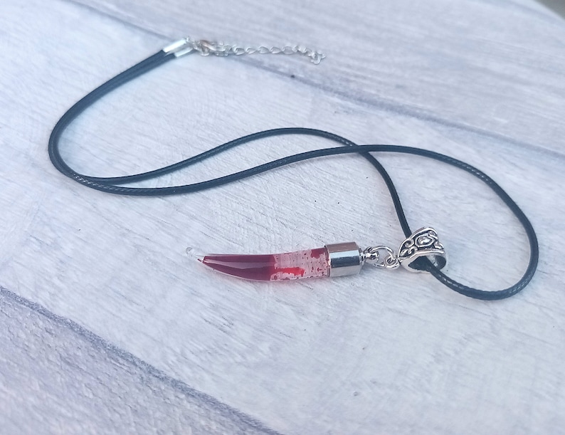 May include: A pendant necklace featuring a clear, horn-shaped charm filled with red liquid and small white particles. The charm is attached to a black cord with a silver-colored clasp and decorative bail.
