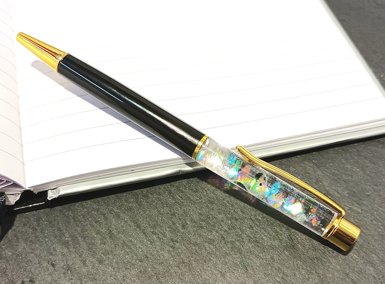 May include: A black and gold pen with a clear barrel filled with colorful glitter. The pen is lying on a white notebook.