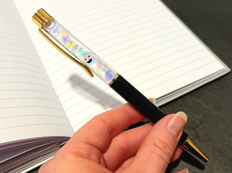 May include: A black and gold pen with a clear barrel filled with iridescent glitter and a small cartoon face. The pen is being held by a hand.