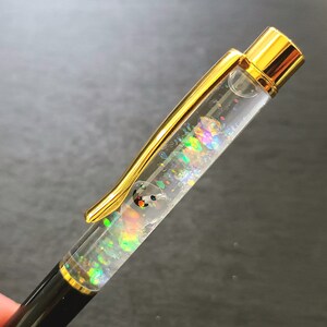 May include: A gold and black pen with a clear barrel filled with iridescent glitter. The glitter is moving inside the pen.