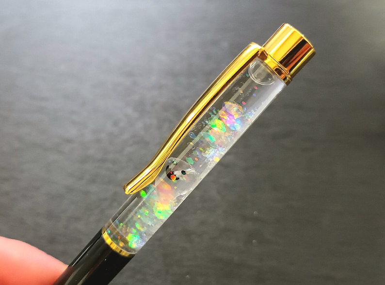 May include: A gold and black pen with a clear barrel filled with iridescent glitter. The glitter is moving inside the pen.