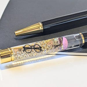 May include: Two black pens with gold accents. One pen has a clear barrel filled with small, clear beads, a pink shape, and a pair of black glasses. The other pen has a gold tip. Both pens are on a dark gray surface.
