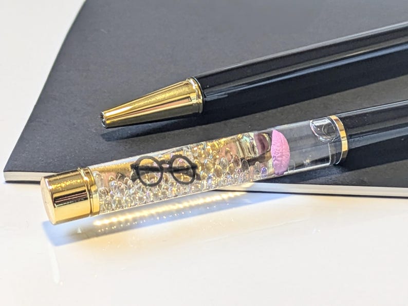 May include: Two black pens with gold accents. One pen has a clear barrel filled with small, clear beads, a pink shape, and a pair of black glasses. The other pen has a gold tip. Both pens are on a dark gray surface.