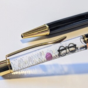 May include: Black and gold pen with a clear barrel filled with small white beads, a purple sphere, and a pair of black glasses. The pen's tip and clip are gold. The pen is lying on a white surface.