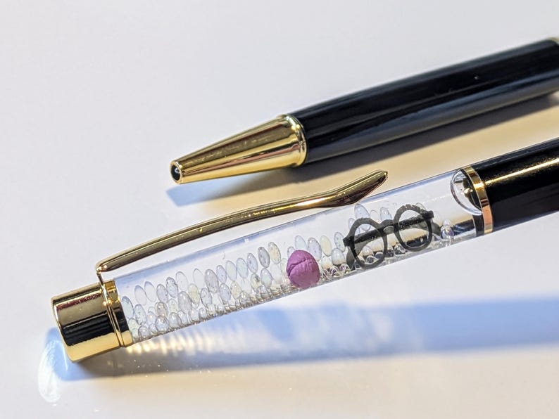 May include: Black and gold pen with a clear barrel filled with small white beads, a purple sphere, and a pair of black glasses. The pen's tip and clip are gold. The pen is lying on a white surface.