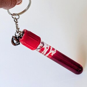 May include: A keychain featuring a miniature, clear vial filled with a deep red liquid and white swirls. The vial has a red cap and is attached to a silver keyring and chain. The keychain is held up against a white background.