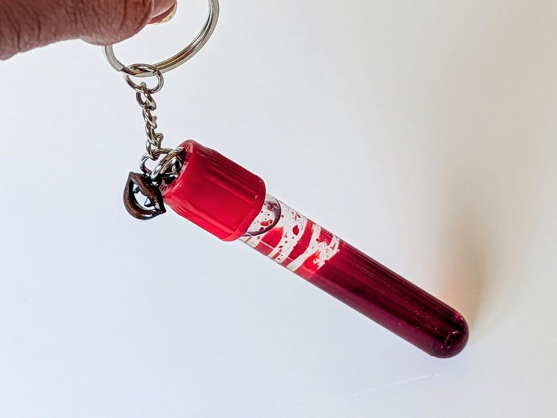 May include: A keychain featuring a miniature, clear vial filled with a deep red liquid and white swirls. The vial has a red cap and is attached to a silver keyring and chain. The keychain is held up against a white background.