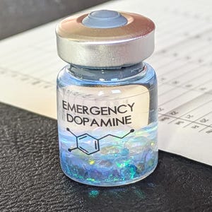 Könnte beinhalten: Ein kleines Glasfläschchen mit der Aufschrift "EMERGENCY DOPAMINE" und einer grafischen Darstellung der Molekülstruktur. Das Fläschchen enthält blaue Flüssigkeit und schillernden Glitzer. Der Verschluss ist silber und blau. Das Fläschchen steht auf einer dunklen Oberfläche neben einem Blatt Papier.